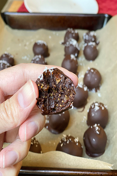 female hand in foreground holding a bitten chocolate truffle and tray of truffles in background