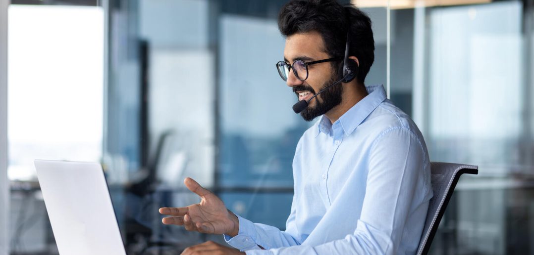Indian young man wearing a headset conducts a webinar, online conference sitting at a laptop in the office.