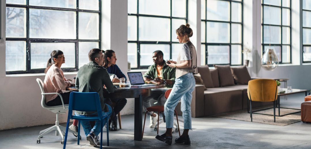 Group of young adults engaged in discussion and work in a bright, stylish co-working space with large windows.