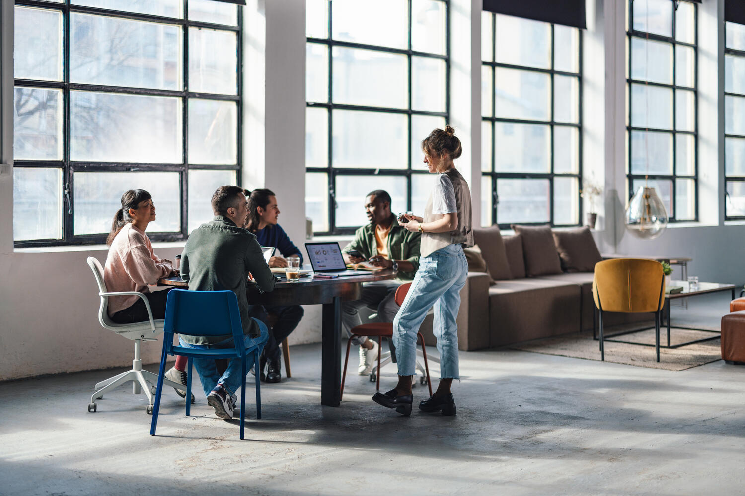 Group of young adults engaged in discussion and work in a bright, stylish co-working space with large windows.