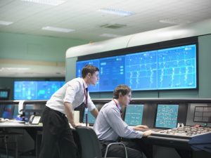 Two men viewing large monitors in a utility control room