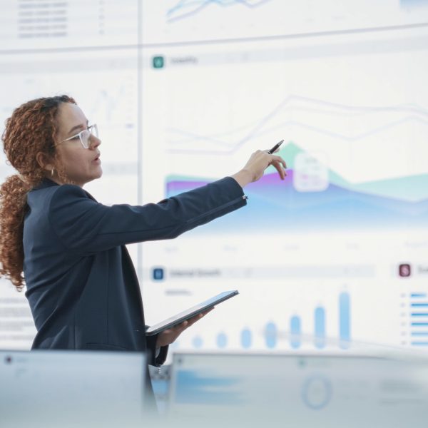 Hispanic Female Senior Data Scientist Reviewing Reports Of Risk Management Department On Big Digital Screen In Monitoring Room. Diverse Consulting Company Employees Working Behind Desktop Computers.