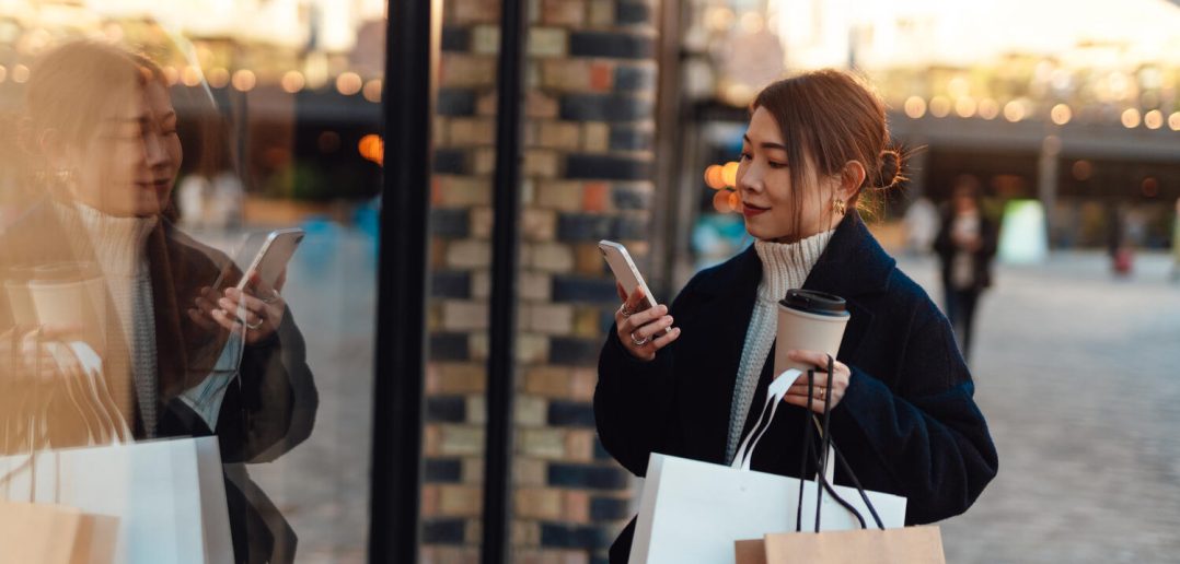 Young woman with shopping bags looking at shop window while using smartphone on the high street in the city. Shopaholic concept.