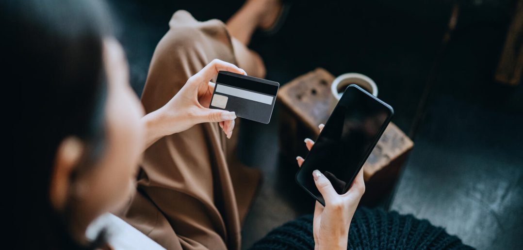 Over the shoulder view of young Asian woman chilling at home, sitting on the floor in bedroom, enjoying a cup of coffee and shopping online with smartphone while making mobile payment with credit card on hand