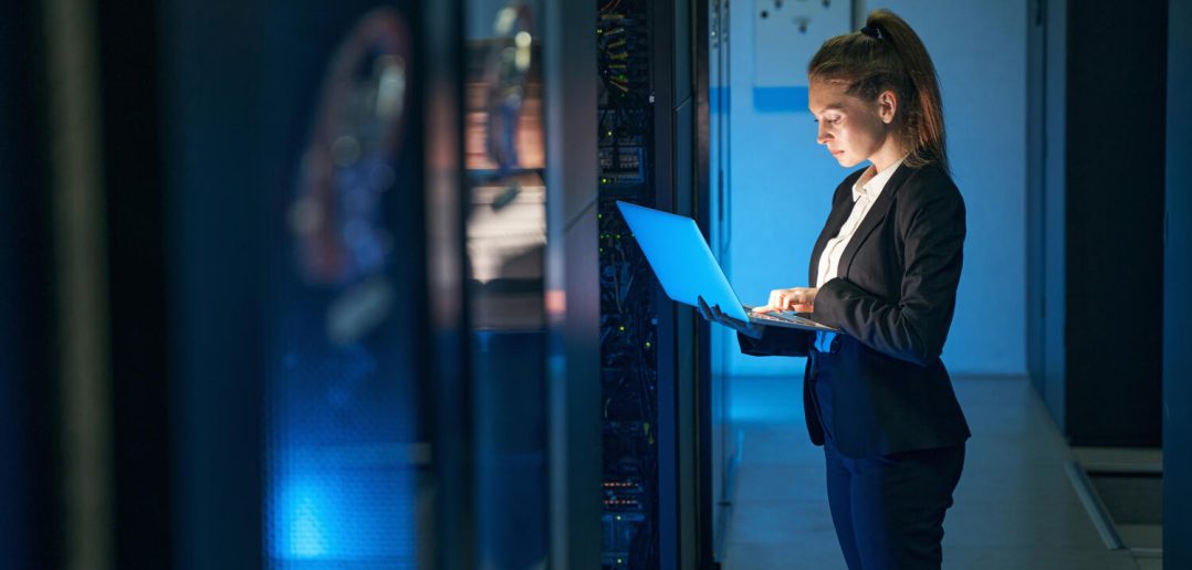 Woman using laptop next to server array in data center.