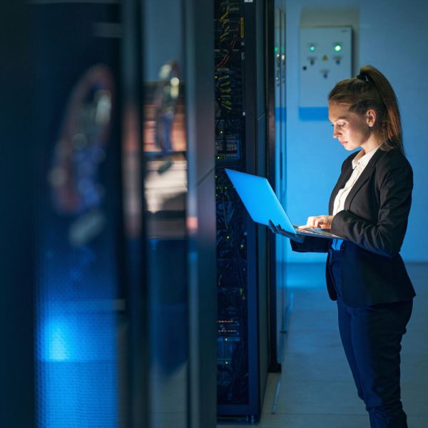 Woman using laptop next to server array in data center.